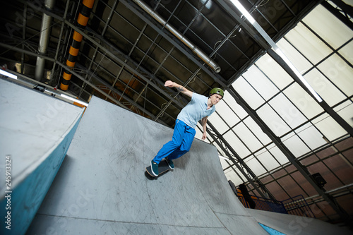 Casual active boy in sports helmet riding down on skateboard while training on special parkour area