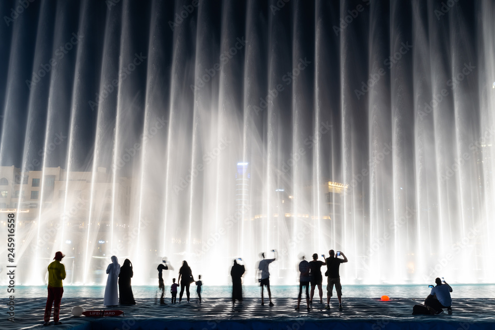 Fototapeta premium Sylwetki ludzi korzystających z pokazu fontann w Dubaju w nocy, Zjednoczone Emiraty Arabskie