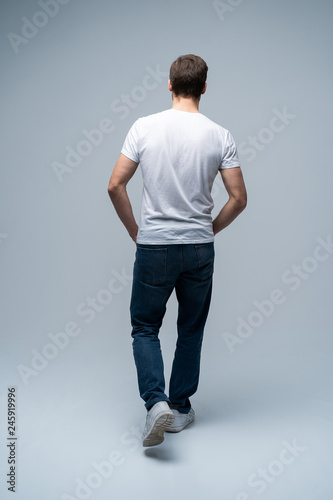 Fotografia back view of a casual young man walking and looking to side on gray background
