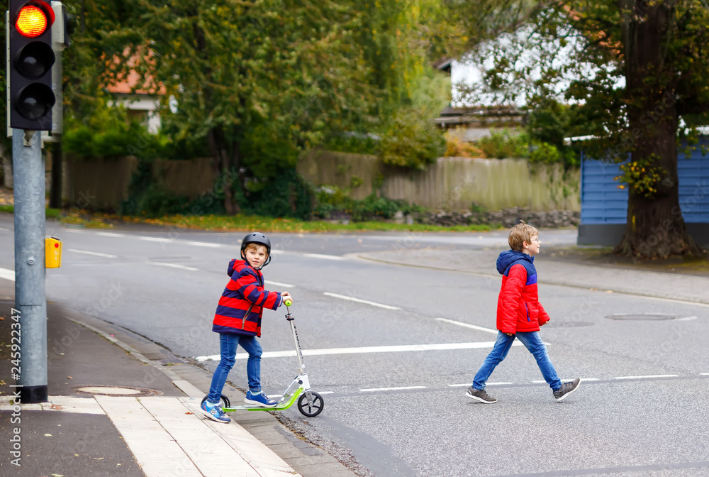 Two little schoolkids boys running and driving on scooter on autumn day. Happy children in colorful clothes and city traffic crossing pedestrian crosswalk with traffic lights.