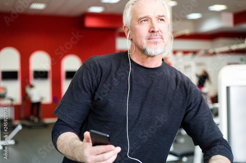 Portrait of a gray-haired male athlete standing in the gym listening to music through headphones photo