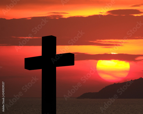 Cross and crucifixion of Jesus Christ silhouette with fiery red dramatic sky and massive glowing large sun over an island during sunset. Religious symbolism, Mourning, memories, remembrance and commem