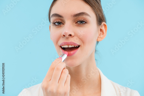 Beautiful young woman posing isolated over blue background holding lip balm.