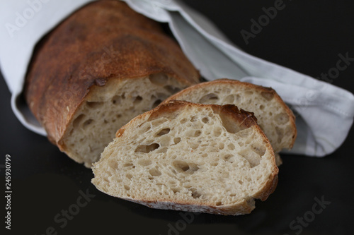 Just baked sourdough loaf - rustic quality homemade wrapped in a napkin with slices of bread lying. Beautiful picturesque close up image with selective focus on black background.