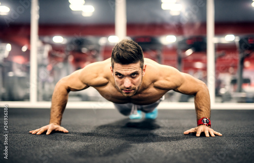 Shirtless Caucasian man doing push ups on gym floor. In background mirror.