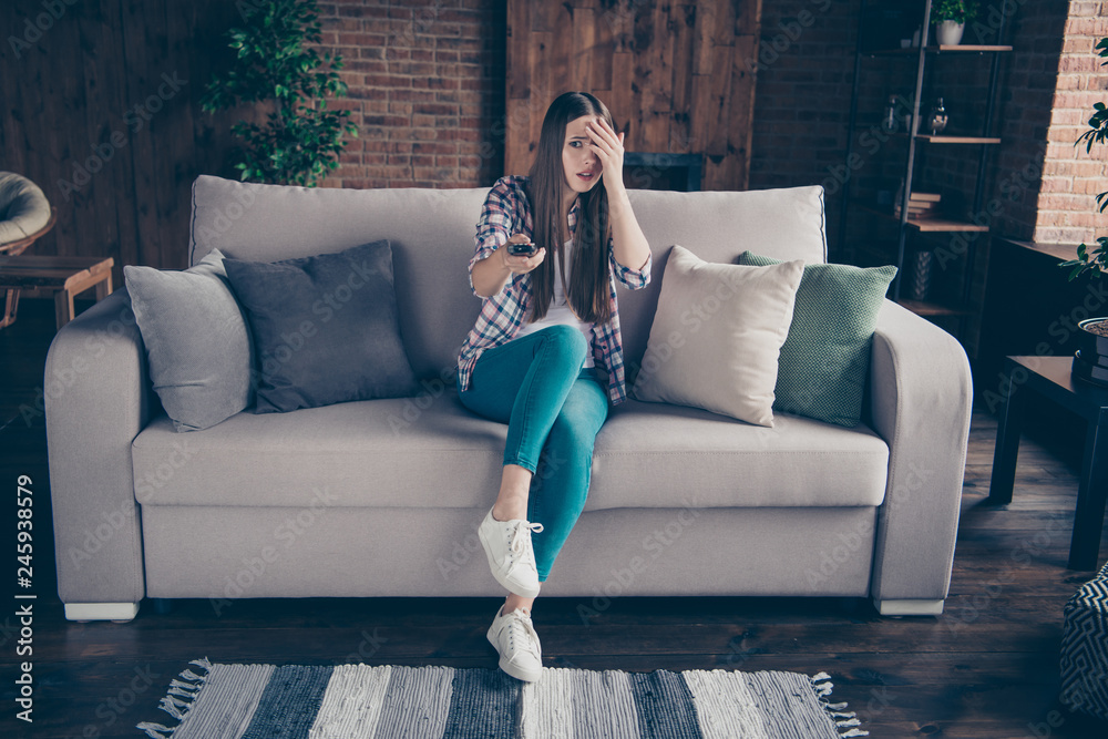 Naklejka premium Full length size photo portrait of scared beautiful curious funky she her lady holding remote control in hand sitting on divan in light loft room