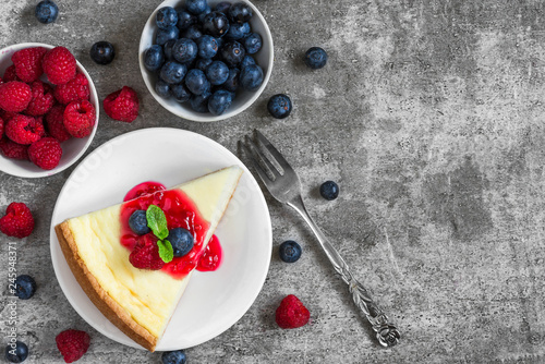 slice of new york cheesecake with fresh raspberries, blueberries, jam and mint with fork on concrete background photo