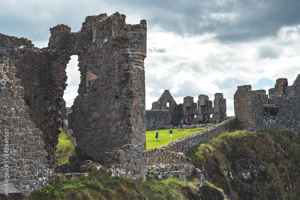Close-up wall of now-ruined Dunluce castle. Northern Ireland. The ruins under the cloudy sky. The tourists visiting the famous archeological site. Historical Medieval building among the wild nature.