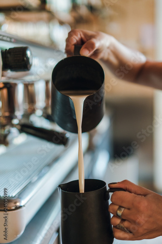 Barista making cafe latte in coffee shop, female hands pouring milk into a black pitchr to steam milk and make cappuccino or latte in a coffee shop