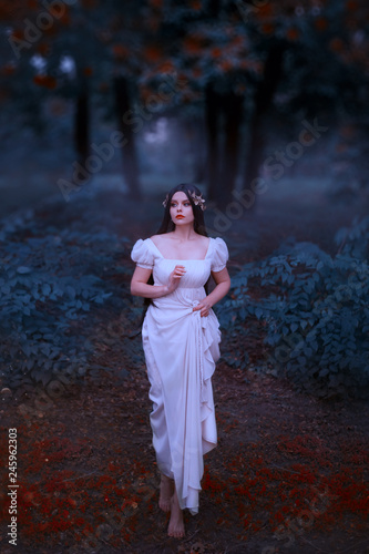 A marvelous, incredible Greek goddess of love, Aphrodite, descended to earth. Young woman with long dark hair in a white dress cautiously walks in the forest. Creative colors, art photography