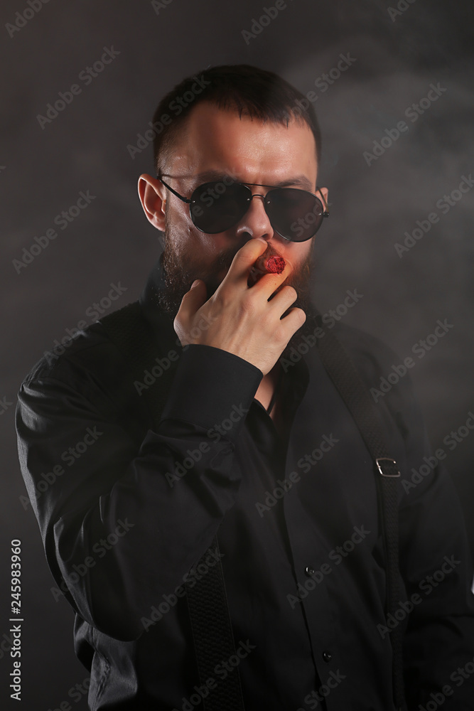 Obraz premium young handsome man with big beard and black sunglasses on black background smoking cigar