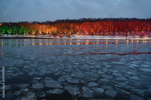 Ice drift on the river in the dusk with colourfully illuminated park in the background. City landscape. Copy space.