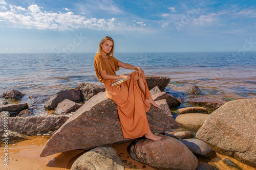 Young girl with long blond hair in long dress is reading book while sitting on stone on sea shore