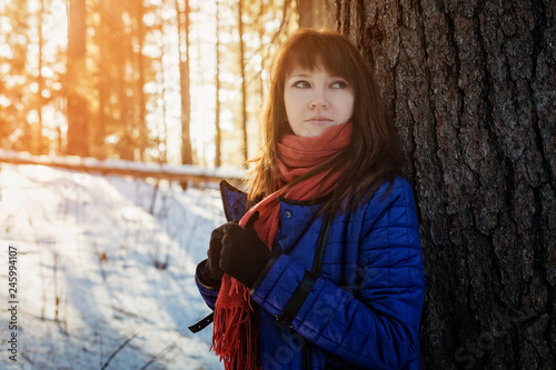 Portrait of a girl in a Sunny winter forest, which stands near a tree in a blue jacket and a red scarf