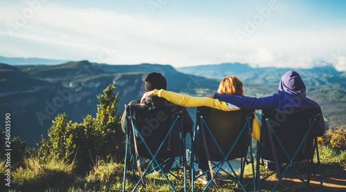 three friends sit in camping chairs on top of a mountain, travelers enjoy nature and cuddle, tourists look into distance on background of panoramic landscape, weekend concept mockup photo