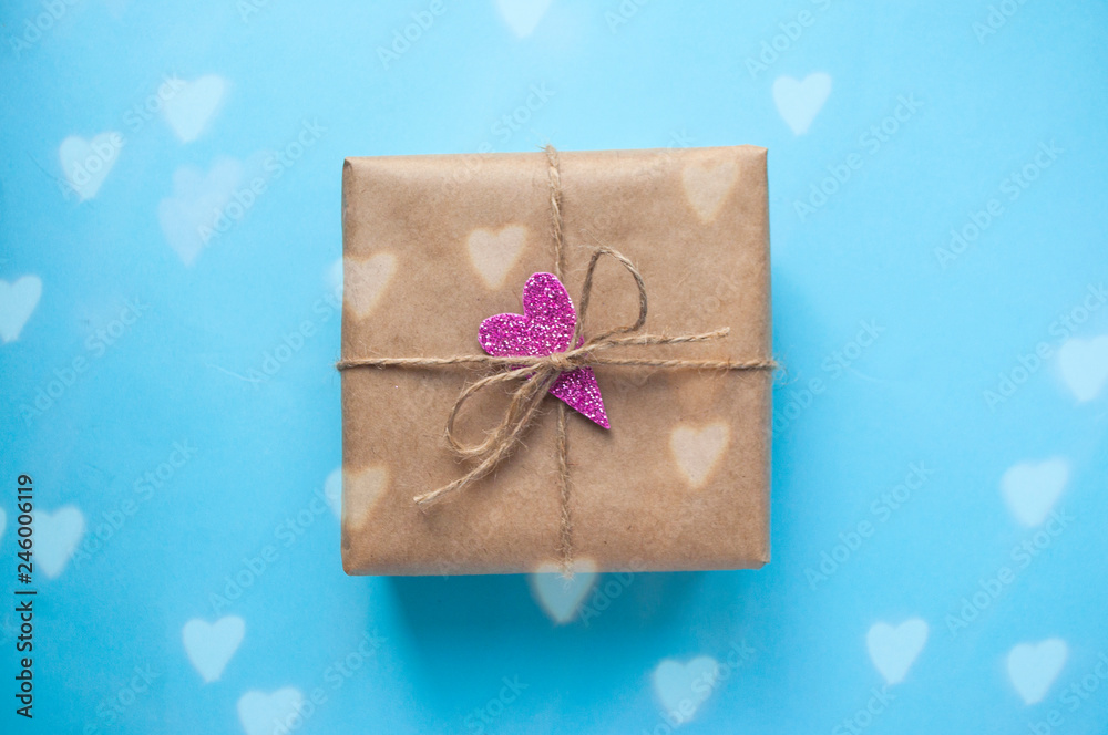 Gift box wrapped in brown colored craft paper with pink glittering heart and tied with rope on blue background. Gift box wrapped in brown colored craft paper with pink glittering heart and tied with rope on blue background.