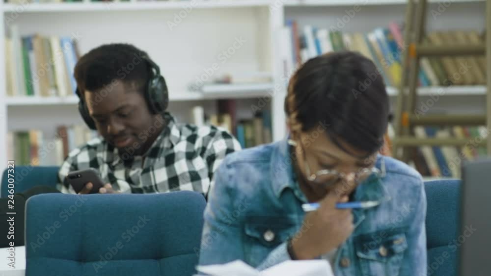 Attractive african female student reading book in the modern library while african male student using smart phone on the background. Attractive african female student reading book in the modern library while african male student using smart phone on the background.