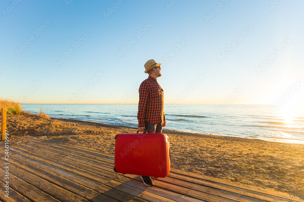 Fototapeta premium Travelling, holidays and people concept - happy man in sunglasses and hat carring his red suitcase near the sea