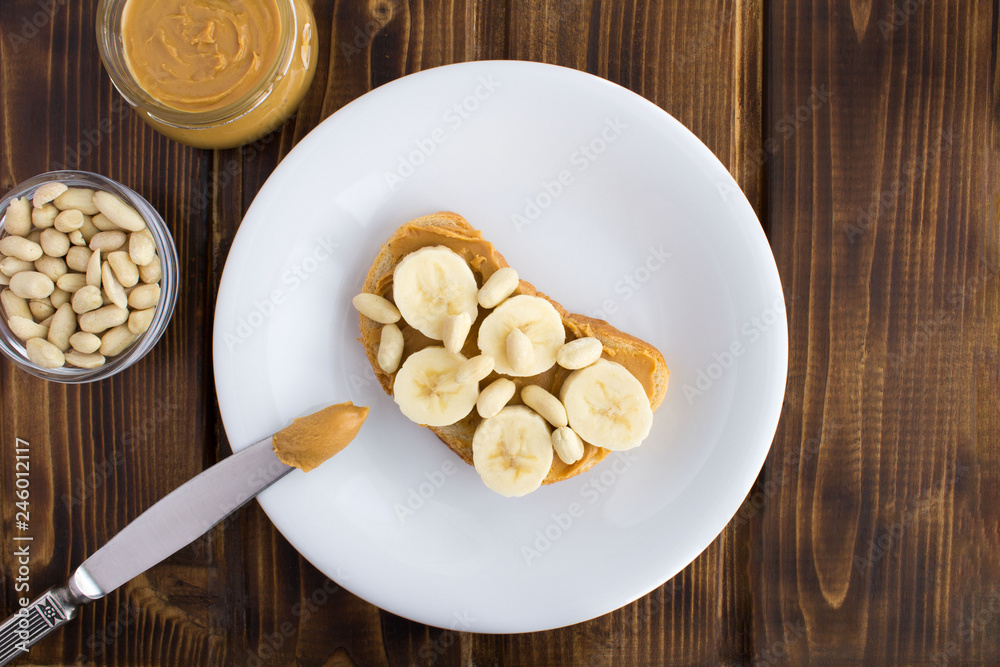 Sandwich with peanut paste and banana in the white plate on the brown wooden background.Top view. Sandwich with peanut paste and banana in the white plate on the brown wooden background.Top view.