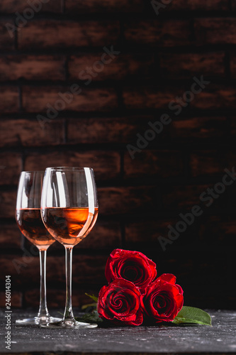 Two wine glasses of rose wine on brick background, bouquet of red roses for romantic evening for Valentines day surprise, marriage proposal passion and love celebration, copy space 