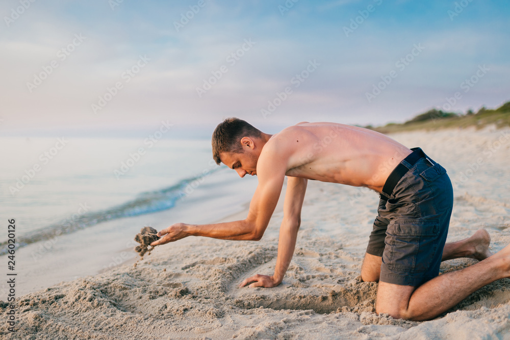 Young topless barefoot man in shorts digging hole by hands on beach beyond sea in summer evening. Funny boy playing on beach with sand on desolate cost near ocean ike child. Vacation leisure and hobby Young topless barefoot man in shorts digging hole by hands on beach beyond sea in summer evening. Funny boy playing on beach with sand on desolate cost near ocean ike child. Vacation leisure and hobby