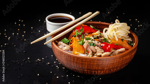 Japanese or Chinese udon noodles with chicken and vegetables, balkar pepper, carrots, parsley, white and black sesame, red onion. noodles in a clay red plate, next to soy sauce and sticks photo
