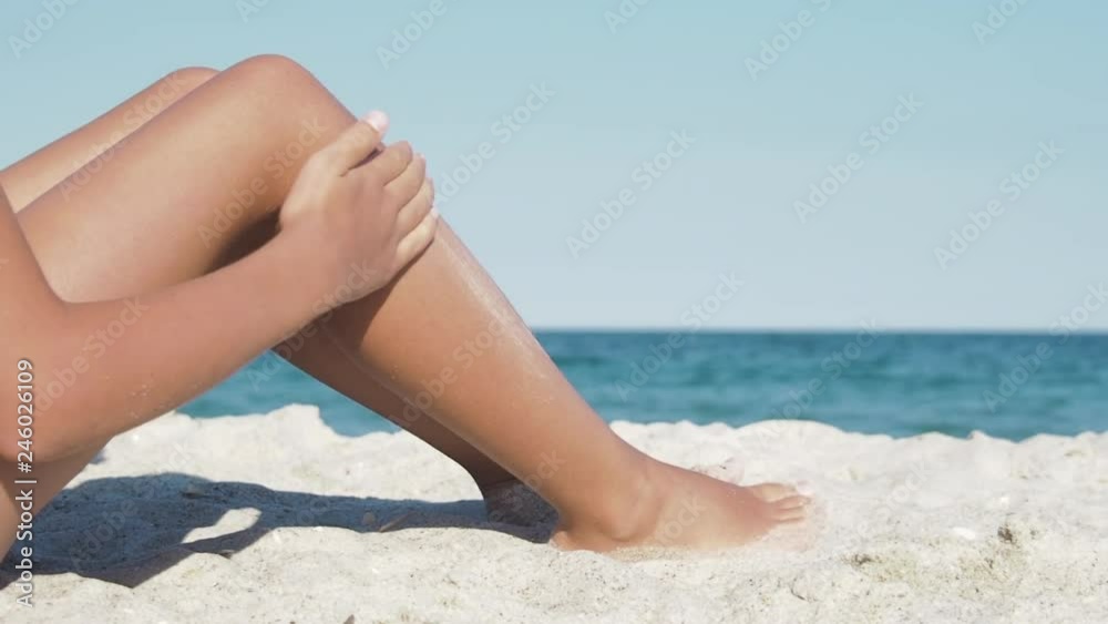 Woman smears sunscree on legs. Legs of a girl sitting on the sand against the background of the sea. Woman smears sunscree on legs. Legs of a girl sitting on the sand against the background of the sea.