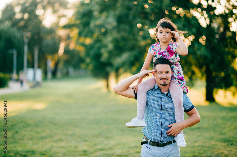 Obraz premium Lifestyle sunset portrait of unusual couple of dad with daughter on shoulders making funny expressive faces. Happy family playing at summer nature outdoor. Fatherhood and childhood. Dad loves his girl