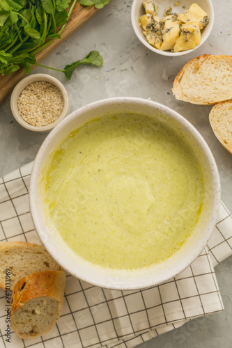 Fresh homemade healthy broccoli cream soup in white bowl with greens, bread and cheese on grey background. top view. vertical