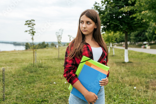 Teenager girl in the summer in the park, in a red shirt in her hands holding notebooks and notes to the school.