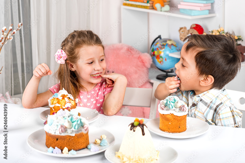 Studio shot of small children, girl and boy, sitting at a table with Easter cakes.  They  eat Easter with festive mood