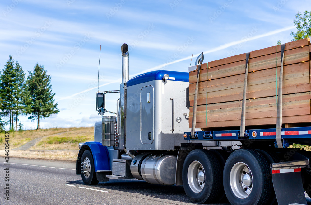 Big rig classic blue bonnet semi truck tractor transporting lumber wood on flat bed semi trailer on the road with hill on the side