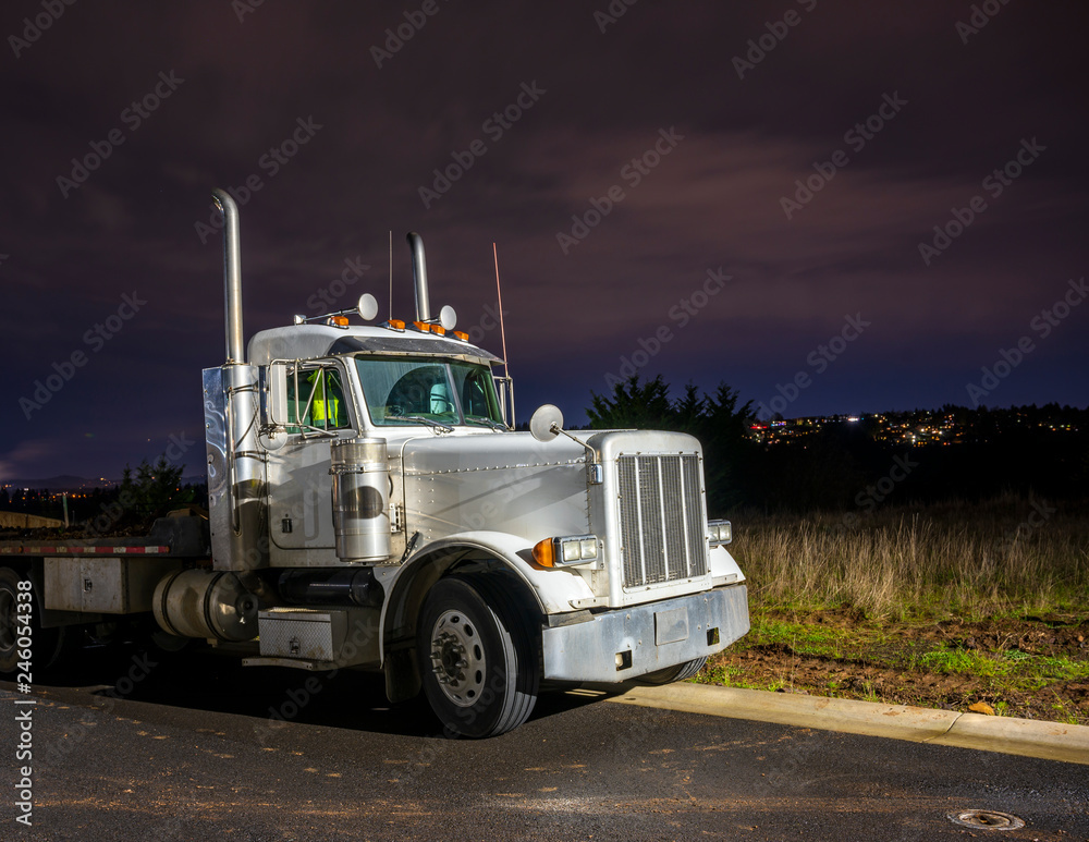 Big rig day cab semi truck with flat bed semi trailer standing on the parking lot in dark night time