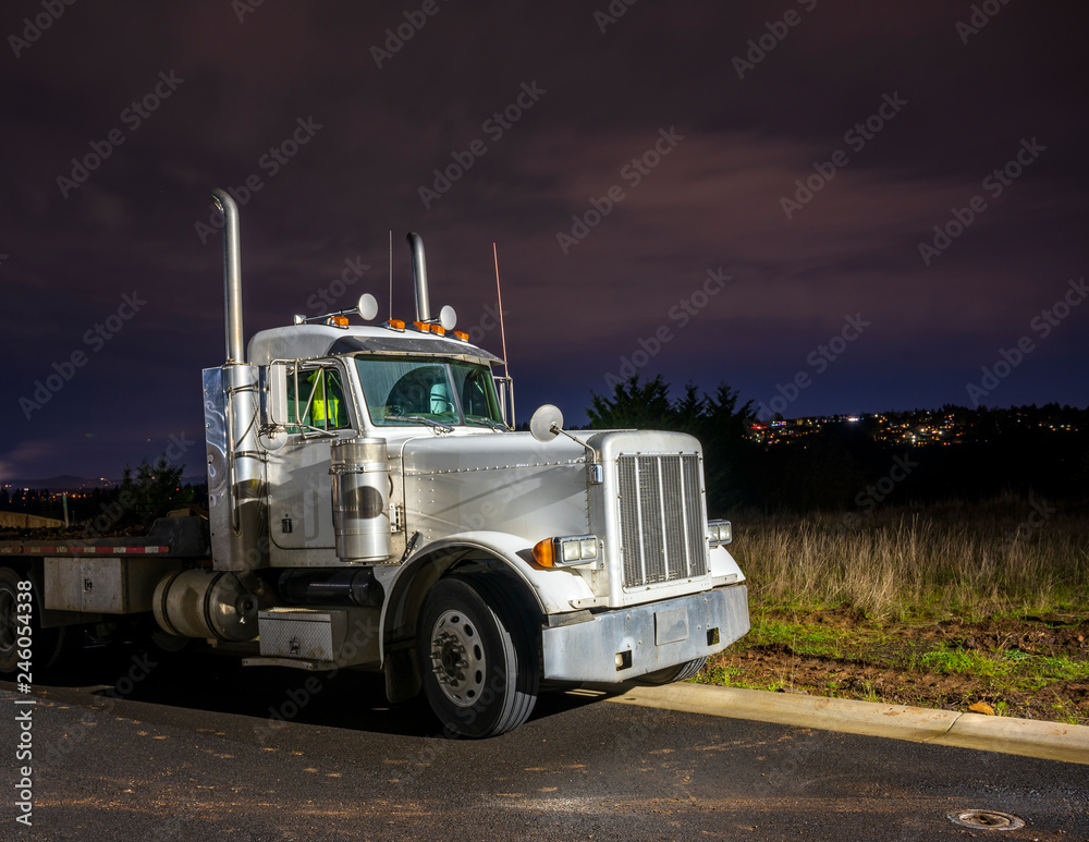 Big rig day cab semi truck with flat bed semi trailer standing on the parking lot in dark night time Big rig day cab semi truck with flat bed semi trailer standing on the parking lot in dark night time