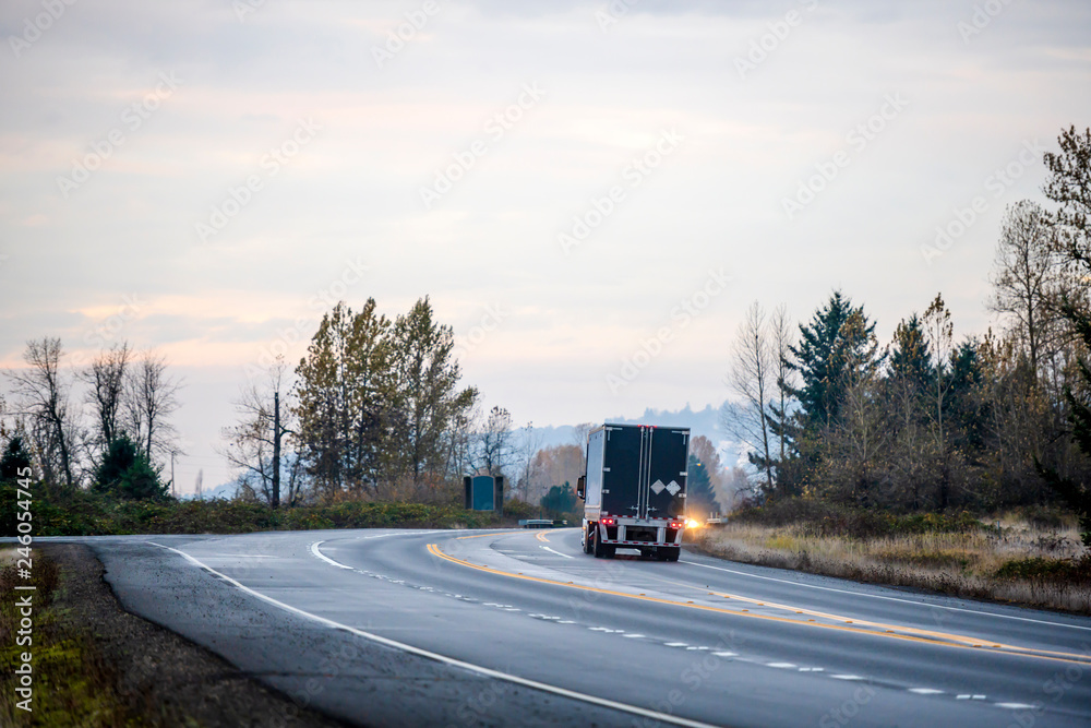 Big rig semi truck with black semi trailer driving on winding wet road in twilight evening time