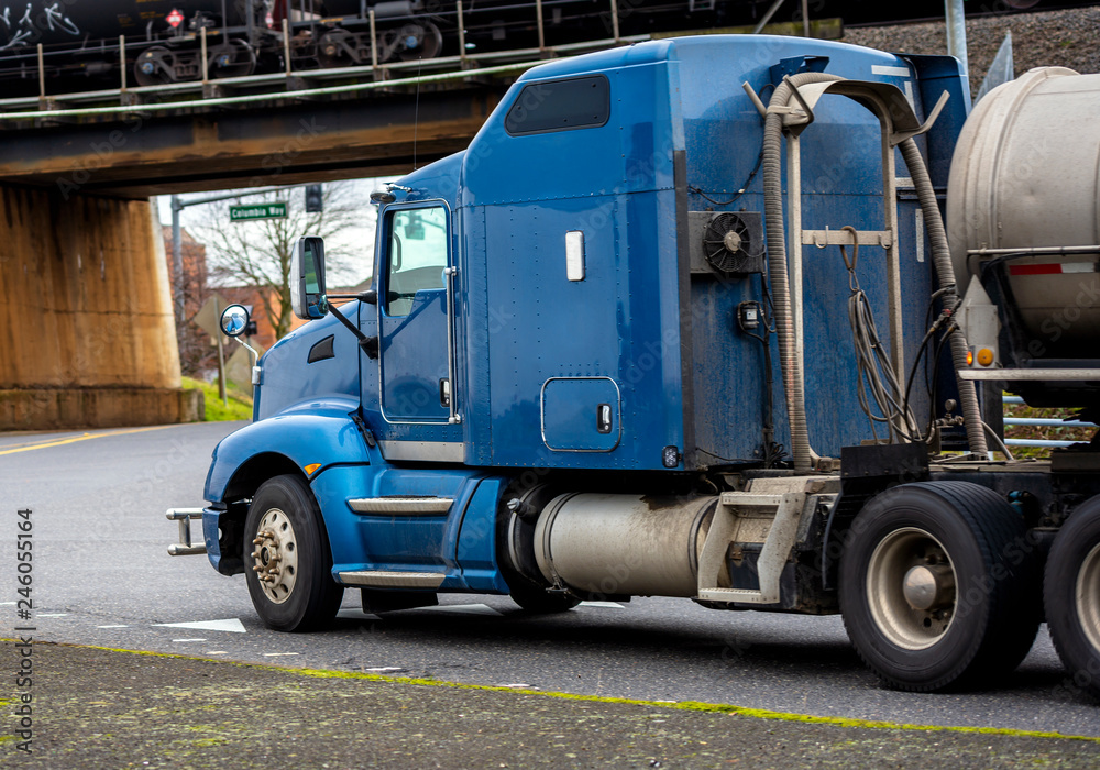 Blue American bonnet rig semi truck with tank semi trailer for transportation of liquids and chemicals running on the road under the bridge