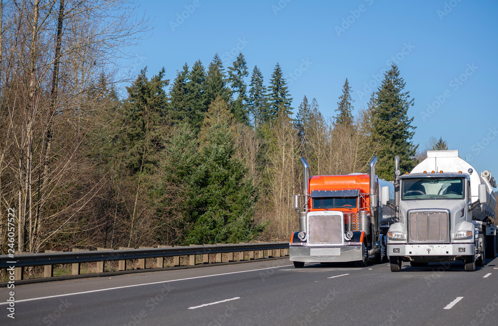 Two big rig orange and white semi trucks with tank semi trailer transporting liquid on the wide road in sunny day