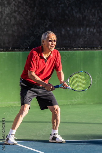 Focused Chinese elderly man ready with racquet raised and serious expression during a game of tennis.
