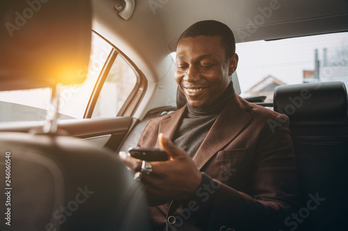 Young smiling african man using smartphone while sitting on backseat in car. Concept of happy business people traveling. © anatoliycherkas