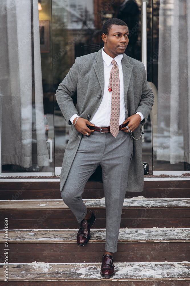 Handsome young african american businessman in classic grey suit holding a smartphone and smiling while leaving the office building Handsome young african american businessman in classic grey suit holding a smartphone and smiling while leaving the office building