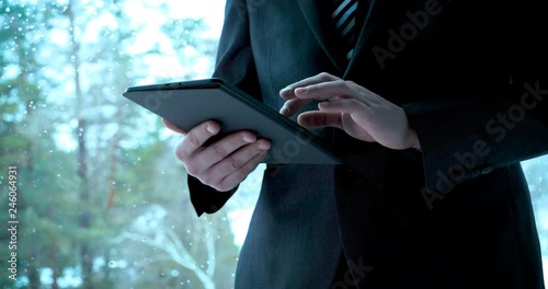 A man (guy) in a suit in a office building on the background of the street is holding a tablet. Concept from: Waiting, Meeting, Marketing, Making a meeting, Looking at a tablet, Writing a message.