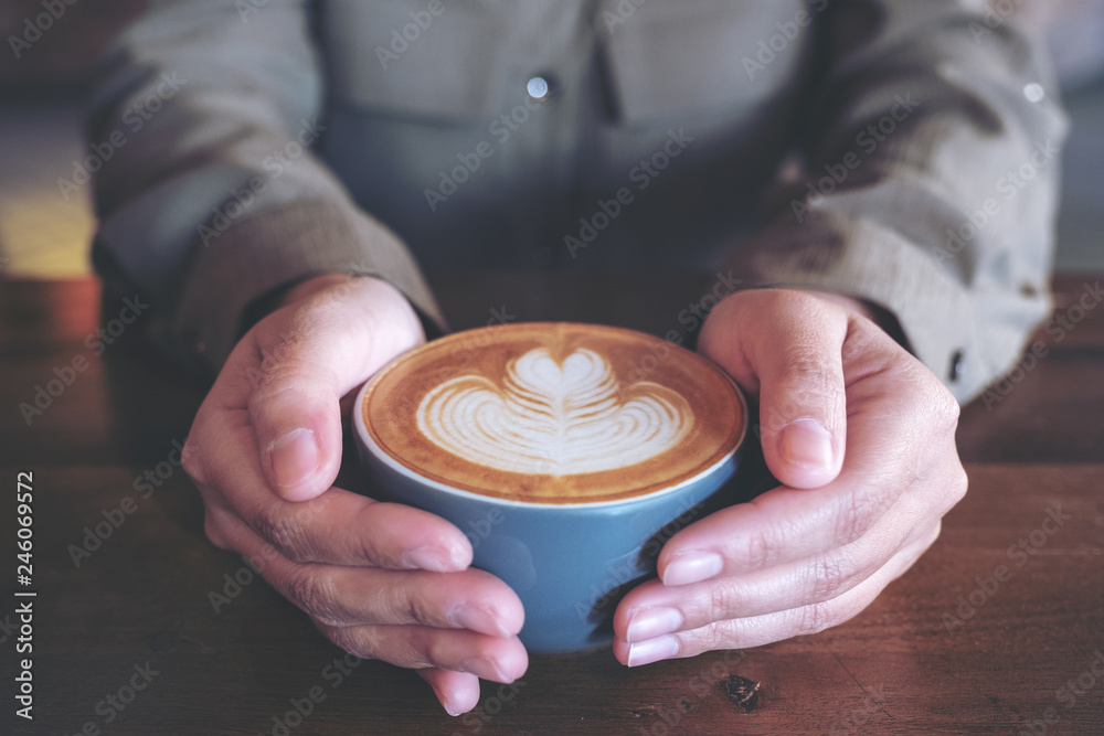 Closeup image of hands holding a blue cup of hot latte coffee with latte art on wooden table in cafe