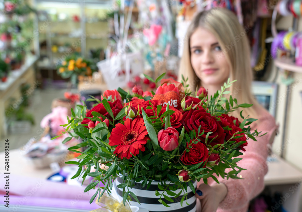 A girl, a florist-designer holds out a beautiful, festive bouquet of red flowers and a heart with the inscription love, specially made for Valentine's day, on the background of a flower shop A girl, a florist-designer holds out a beautiful, festive bouquet of red flowers and a heart with the inscription love, specially made for Valentine's day, on the background of a flower shop