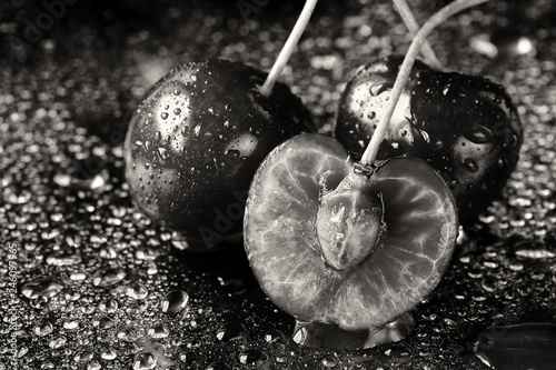 Monochrome artistic extreme close up of two and a half sliced cherry fruits with the stems and pits. Black background with water drops and reflections