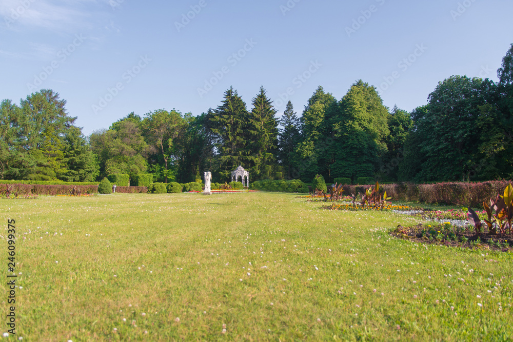 a large green trimmed lawn in a park with statues and a gazebo, with trees and shrubs around