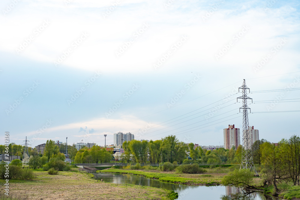 a small river in a park with green trees and bushes on the background of multi-storey houses and blue sky with white clouds