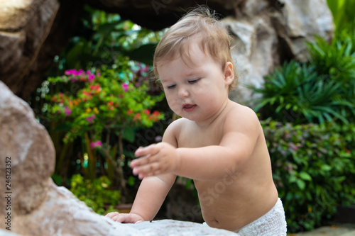 Pretty crawling baby girl smiling and crawling on the wooden brown walkway on the background of green bushes and trees. The child found on the floor and trying to eat. There is a place for text.