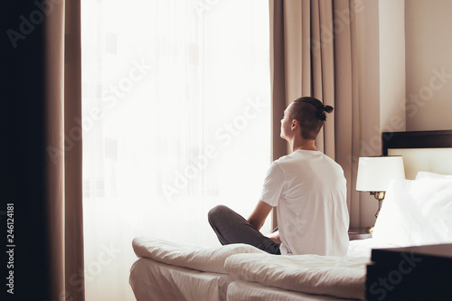 Cute young guy woke up and meditates on his bed in the lotus position looking out the window of his bedroom. Concept of cheerfulness and good mood for the whole day