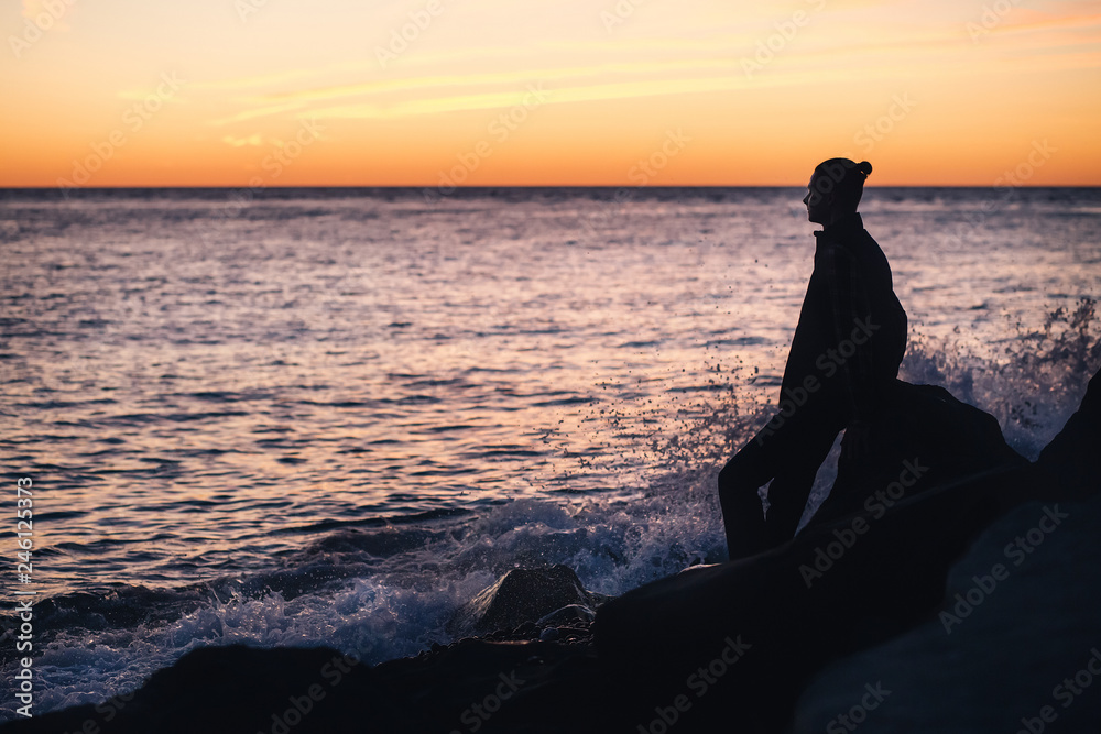 Silhouette of a slim young man leaning on a stone on the coast and looking at the sea surface on a warm summer evening. Romantic holiday concept. Copyspace Silhouette of a slim young man leaning on a stone on the coast and looking at the sea surface on a warm summer evening. Romantic holiday concept. Copyspace