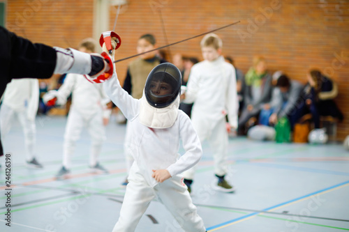 Little kid boy fencing on a fence competition. Child in white fencer uniform with mask and sabre. Active kid training with teacher and children. Healthy sports and leisure. © Irina Schmidt